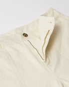Close-up of a cream-colored garment, likely a jacket or vest, showing a buttoned flap, a zippered pocket, and a small brown button detail against a light gray background.