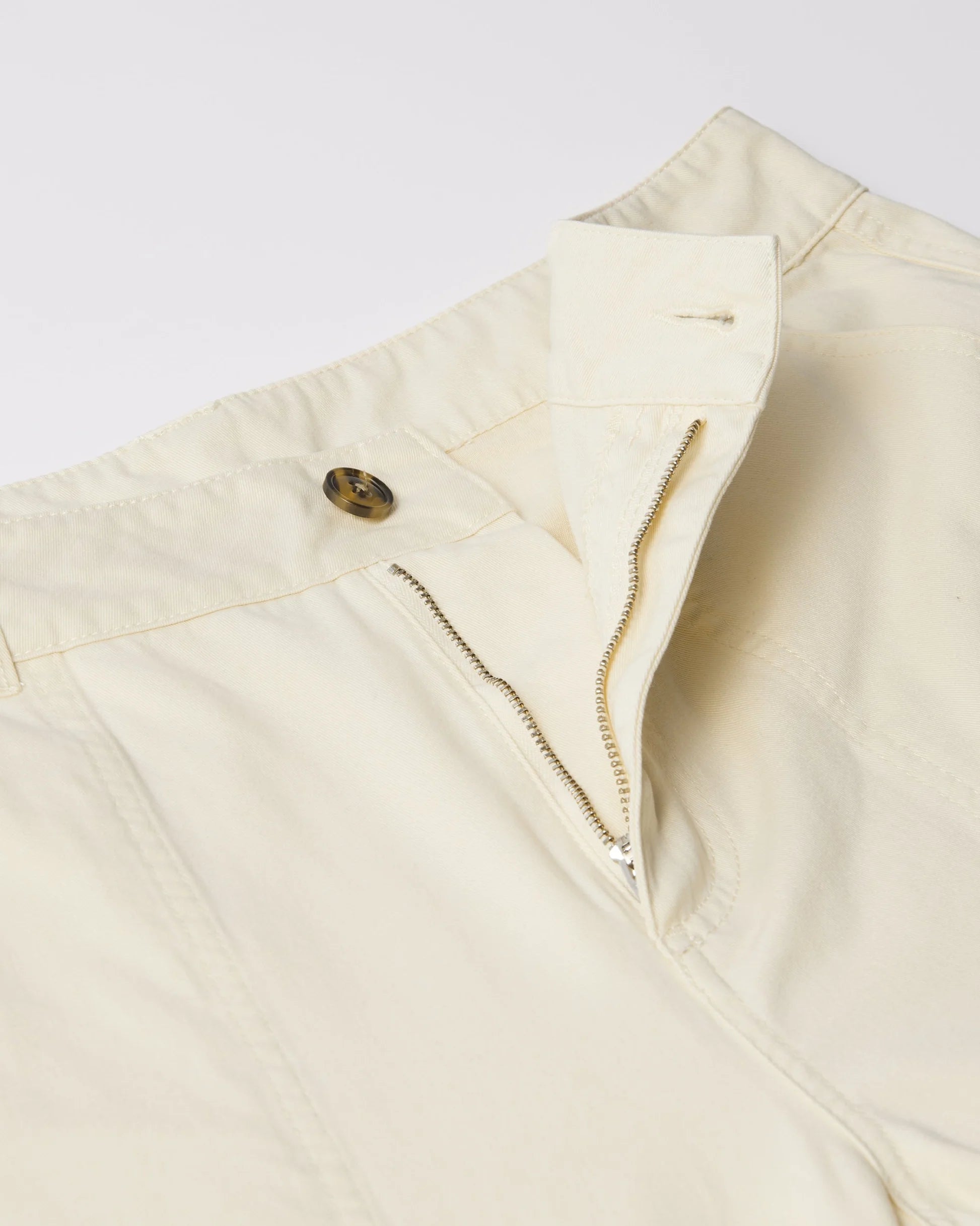 Close-up of a cream-colored garment, likely a jacket or vest, showing a buttoned flap, a zippered pocket, and a small brown button detail against a light gray background.