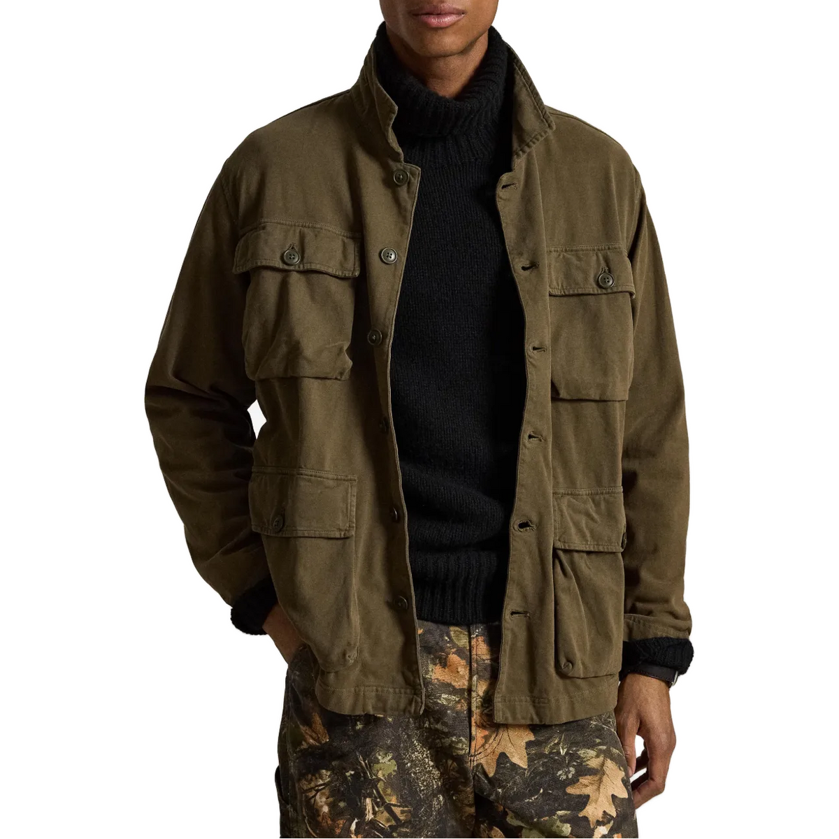 A man wearing an olive green military-style field jacket over a black turtleneck sweater and camouflage pants, shown from the waist up against a plain white background.