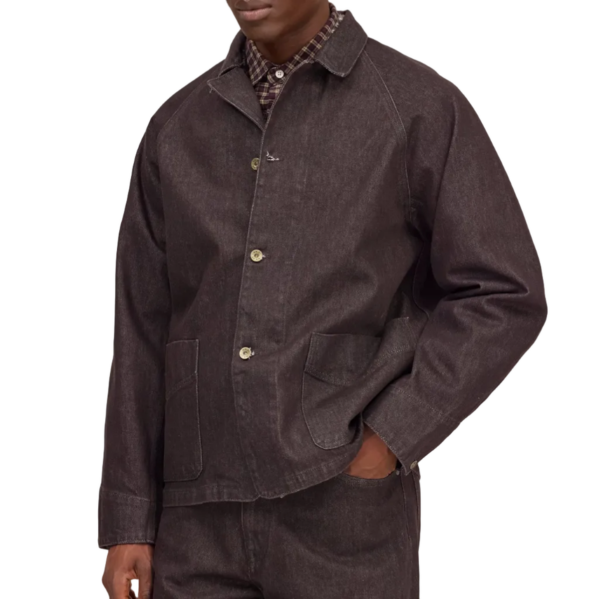 A man wearing a dark brown corduroy utility jacket over a plaid shirt and matching dark brown corduroy trousers, standing against a solid light beige background.