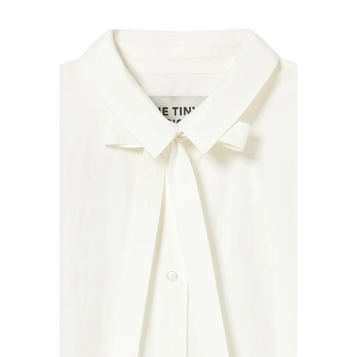 Close-up of the collar on The Tiny Big Sister Bow Neck Shirt, showing the bow at the neck, the brand label and a button on the front.