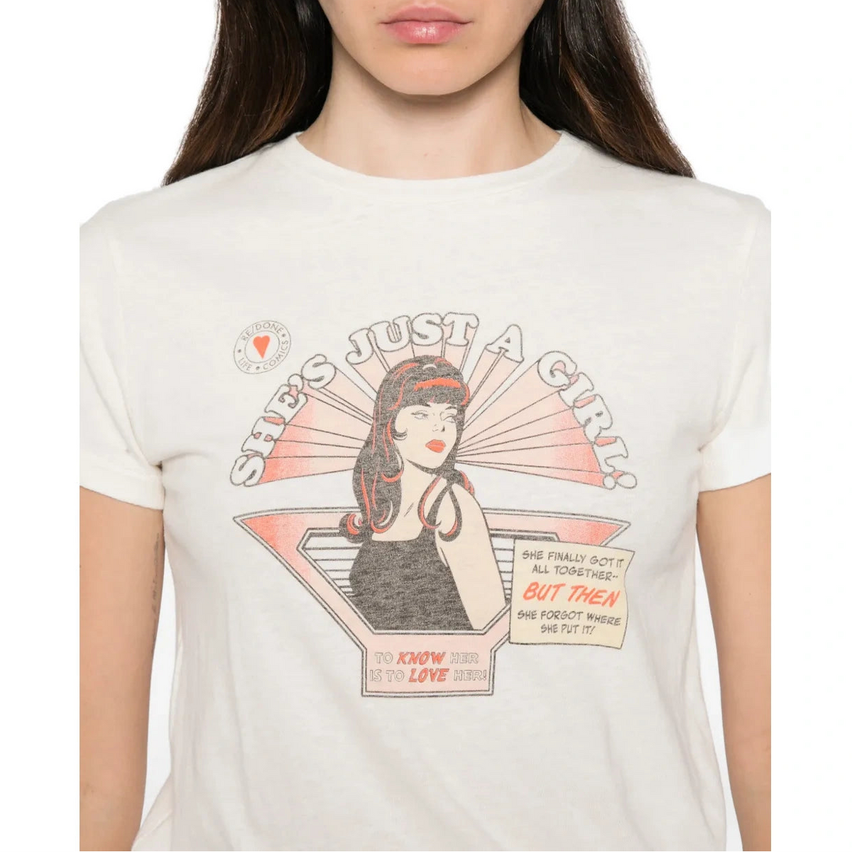 A woman wearing a cream-colored graphic t-shirt with a vintage-style illustration of a girl and the text 'SHE'S JUST A GIRL!'.