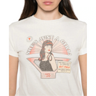 A woman wearing a cream-colored graphic t-shirt with a vintage-style illustration of a girl and the text 'SHE'S JUST A GIRL!'.