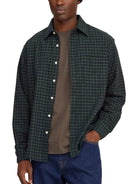 A man wearing a dark green and black plaid button-down shirt over a brown t-shirt and blue jeans, shown from the waist up against a plain white background.