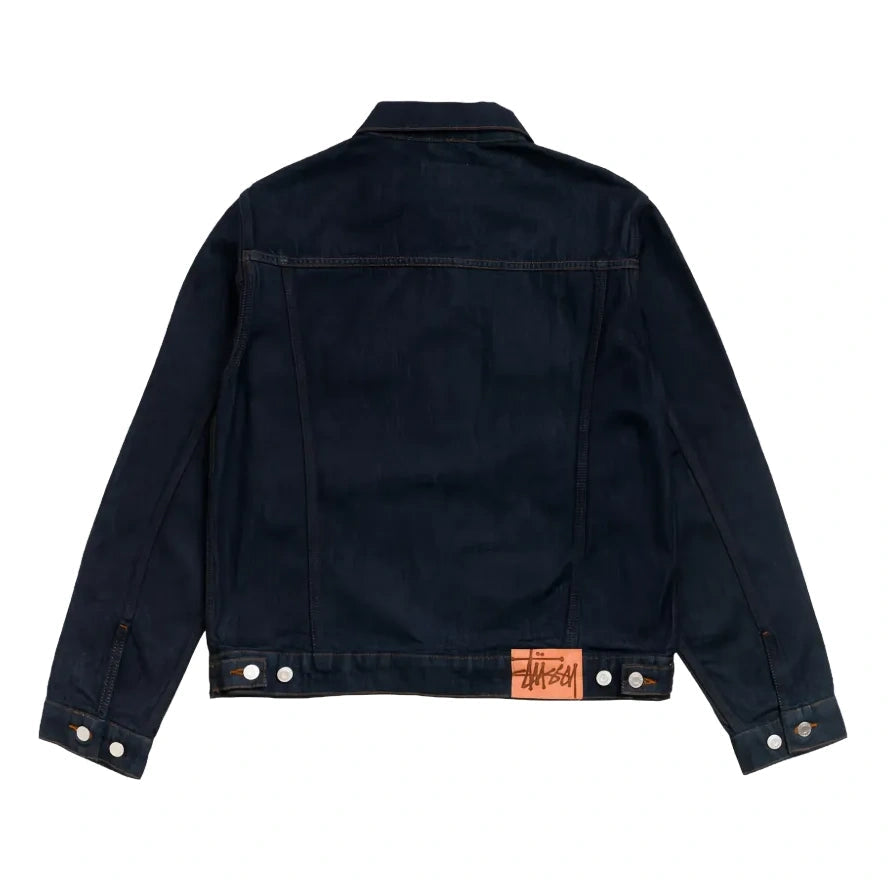 Back view of the rinsed indigo Trucker Denim jacket laid flat, showing the back yoke, button waist tabs and a Stüssy patch at the hem.