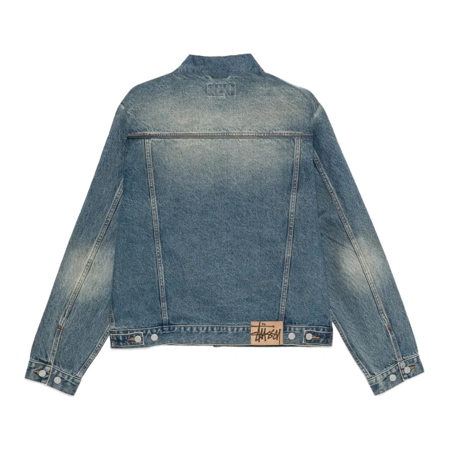 Back view of the dark wash Trucker Denim jacket laid flat, showing fading across the back and sleeves, the button waist tabs and a Stüssy patch at the hem.
