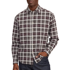 A man wearing a long-sleeve button-down plaid shirt in dark purple , white, and gray, paired with dark blue jeans, standing against a whitee background.