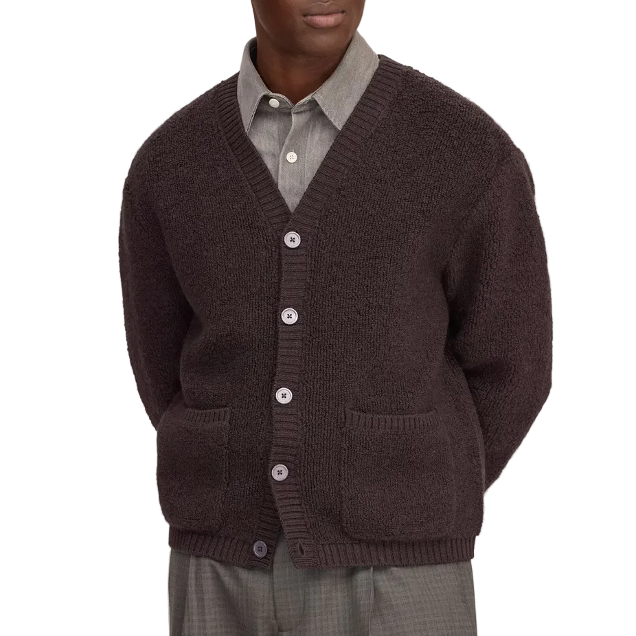A man wearing a dark purple knit cardigan over a gray collared shirt and gray trousers, standing against a white background.