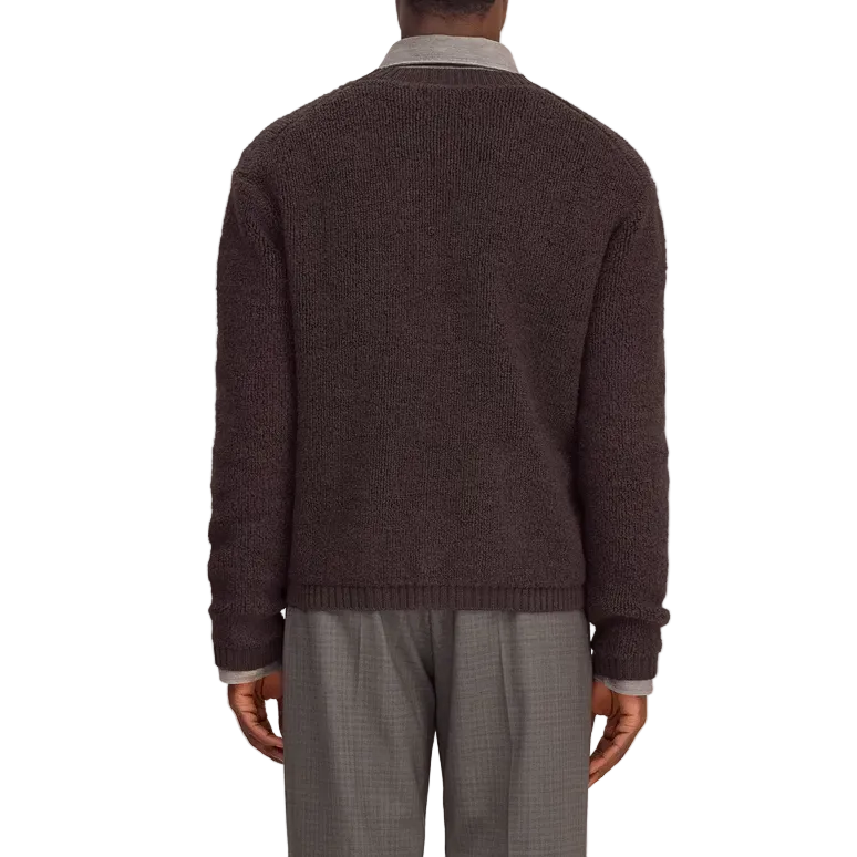 A man viewed from behind wearing a dark purple knit sweater over a light gray collared shirt and gray trousers, standing against a white  background.