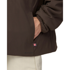 Close-up of a hand in the side pocket of a dark brown Snow Peak Flexible Insulated Cardigan, showing the red PrimaLoft tag at the hem, the tonal Snow Peak logo and the elastic cuff.