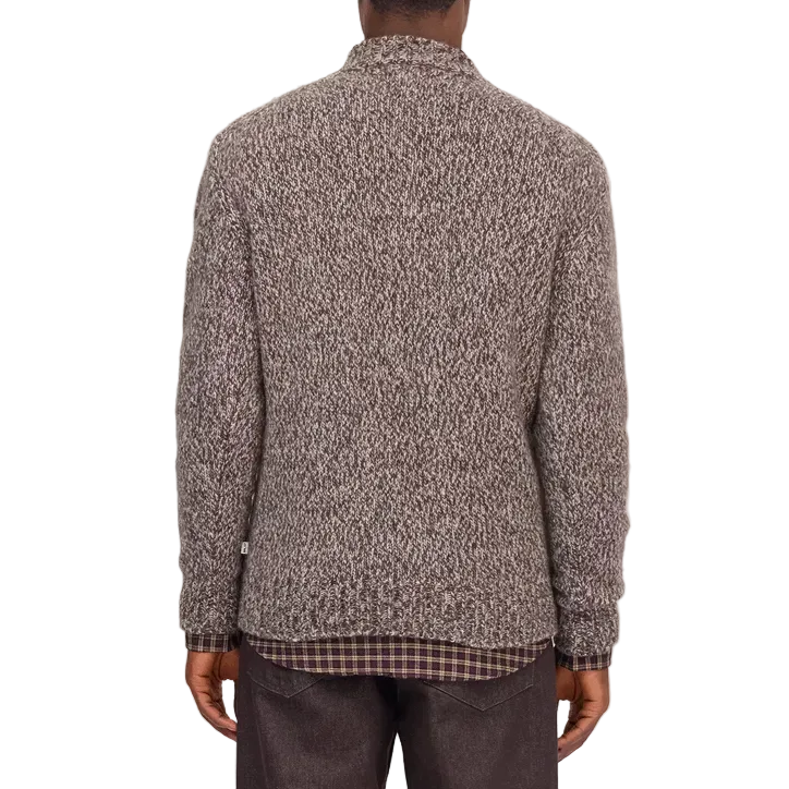 A man viewed from the back wearing a textured gray and black knit mock-neck sweater over a dark plaid shirt and dark brown trousers, set against a white background.