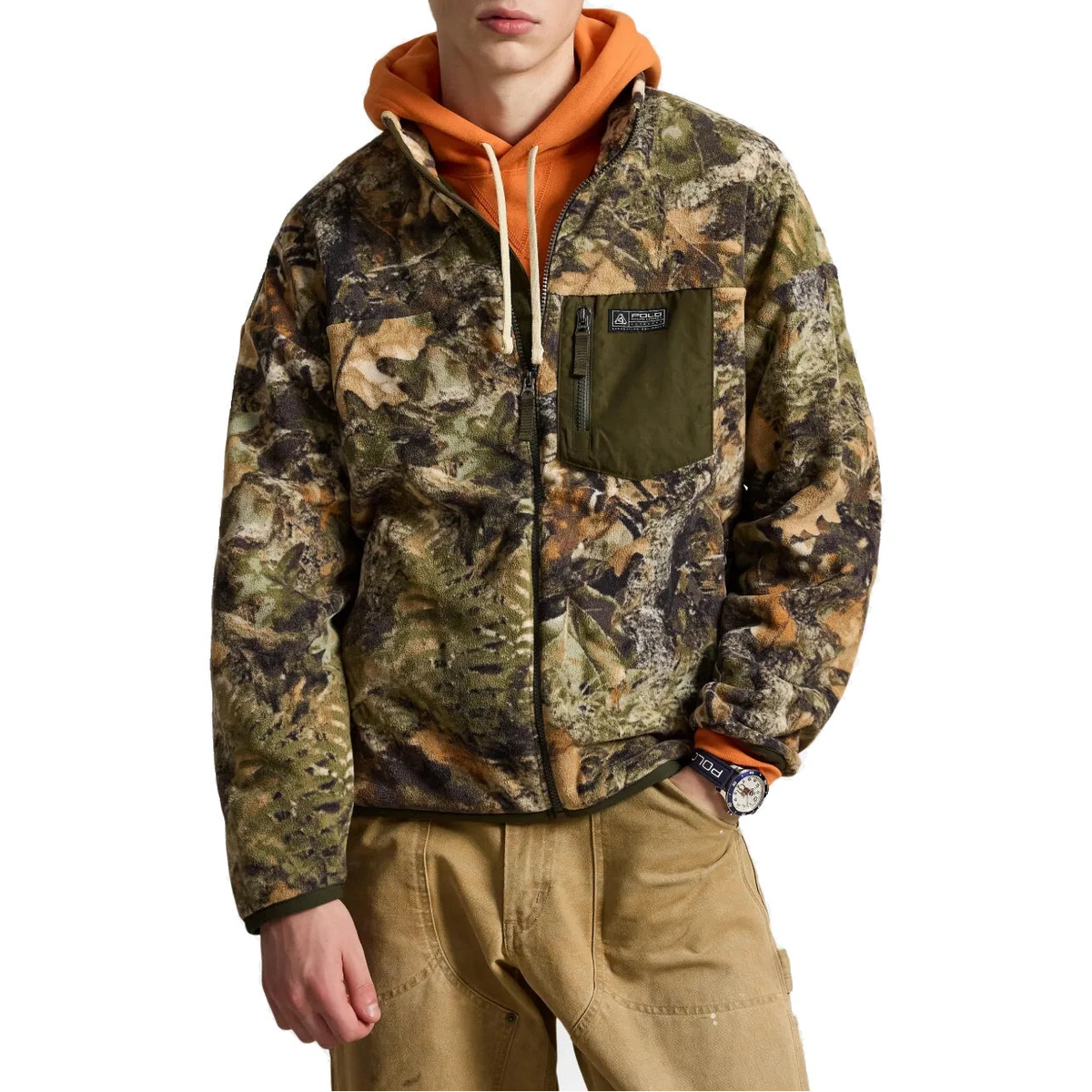 A man wearing a camouflage zip-up jacket over an orange hoodie and tan pants, shown from the waist up against a plain white background.