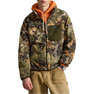 A man wearing a camouflage zip-up jacket over an orange hoodie and tan pants, shown from the waist up against a plain white background.