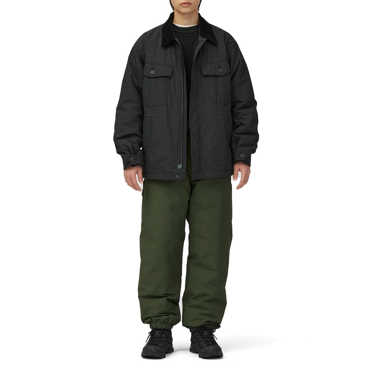 Full-length view of a model wearing the black Snow Peak Hybrid Takibi Adjustable Jacket unzipped over a black knit sweater, with olive green pants and black hiking shoes.