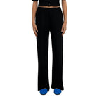 A model stands against a plain white wall wearing a black short-sleeve cropped top with a button front and matching black wide-leg pants, paired with bright blue shoes.