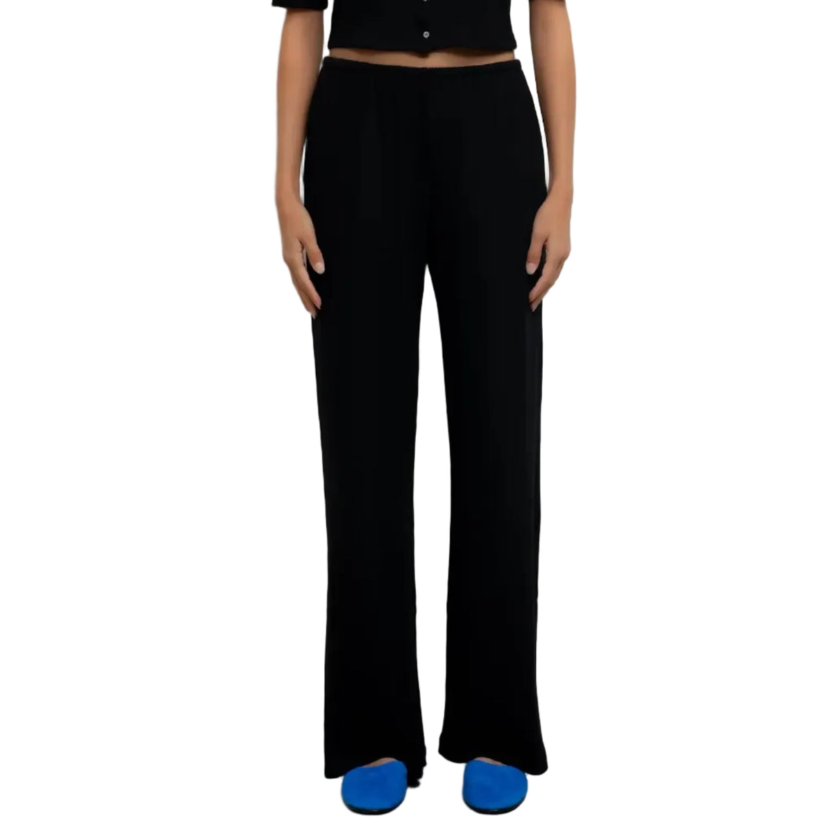 A model stands against a plain white wall wearing a black short-sleeve cropped top with a button front and matching black wide-leg pants, paired with bright blue shoes.