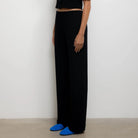 A model stands in profile wearing a black short-sleeve cropped button-front top and matching black wide-leg trousers, with bright blue shoes, against a plain light gray wall.