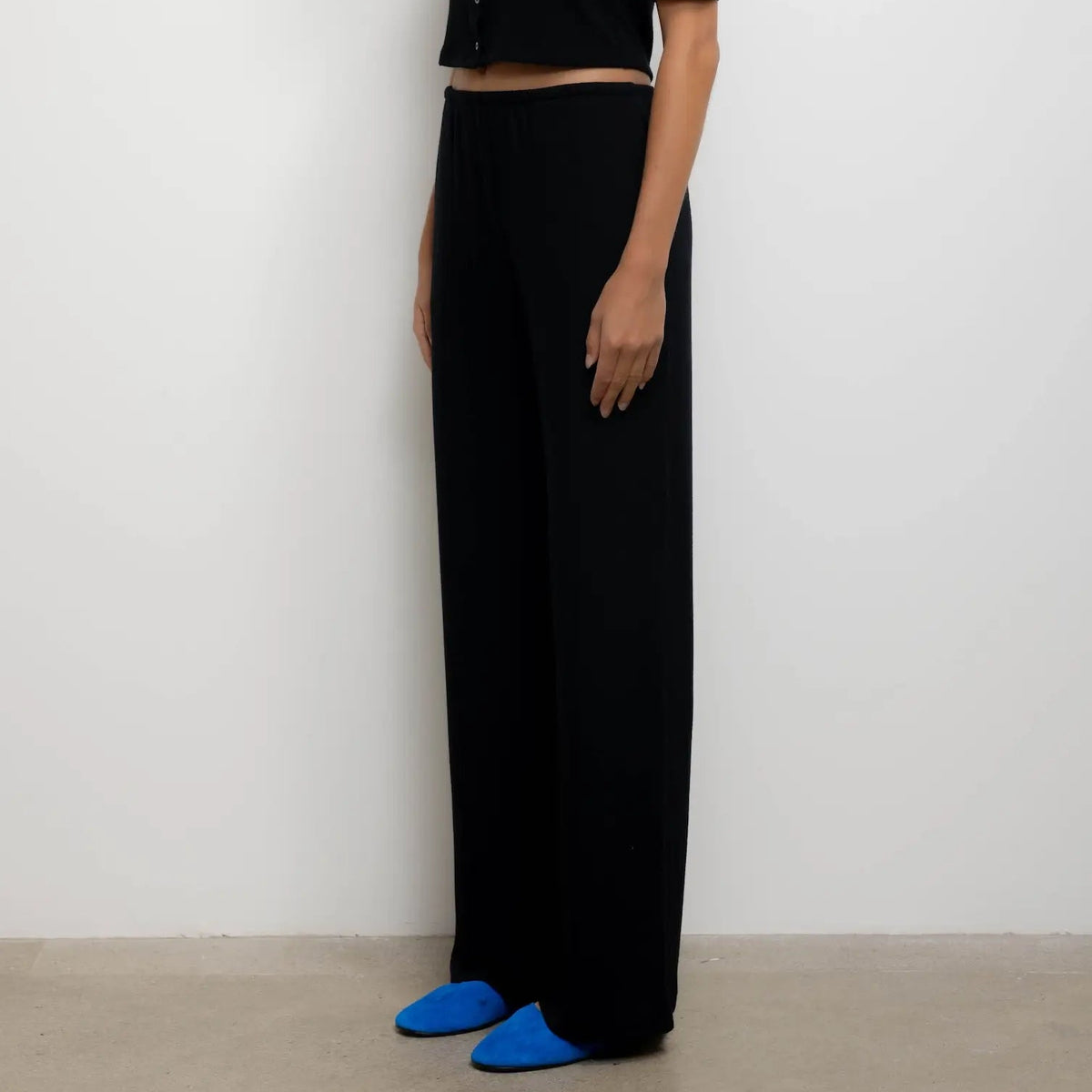 A model stands in profile wearing a black short-sleeve cropped button-front top and matching black wide-leg trousers, with bright blue shoes, against a plain light gray wall.