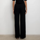 A model viewed from the back wearing a black short-sleeve top and matching black wide-leg trousers, standing against a plain white wall.