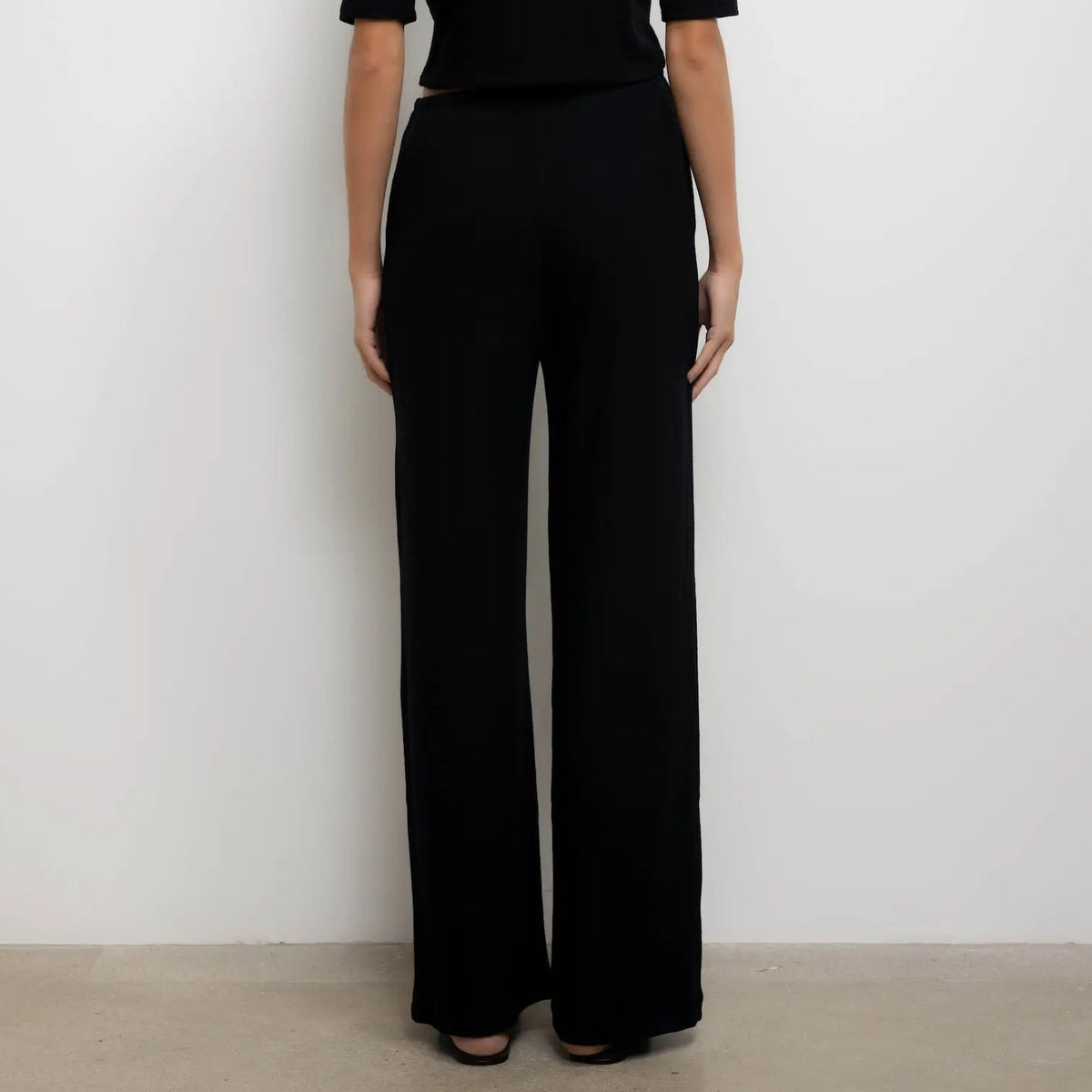 A model viewed from the back wearing a black short-sleeve top and matching black wide-leg trousers, standing against a plain white wall.
