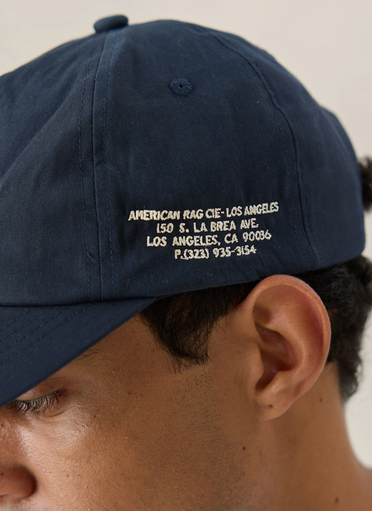Close-up of a person wearing a navy blue baseball cap with white embroidered text on the side reading 'AMERICAN RAG CIE - LOS ANGELES' and an address.