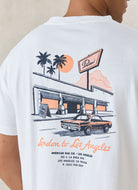 A person wearing a white graphic T-shirt, shown from the back. The shirt features a retro-style illustration of a building with palm trees, a sunset, a vintage car, and orange text reading 'London to Los Angeles' with smaller address text below.