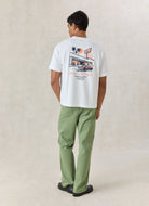 A man stands with his back to the camera wearing a white graphic t-shirt with a retro design on the back, paired with sage green pants and black shoes, against a textured beige studio backdrop.