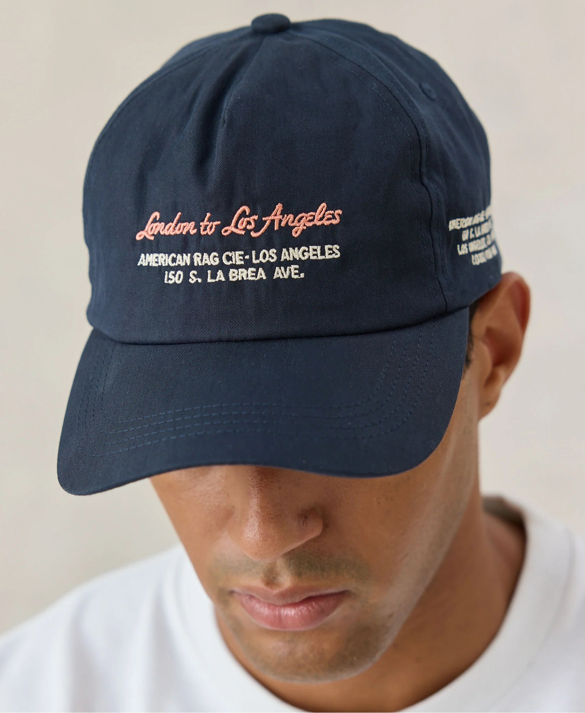 A navy blue baseball cap with embroidered text reading 'London to Los Angeles' and 'American Bag Cie. Los Angeles' on the front, worn by a man in a white t-shirt against a neutral beige background.
