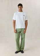 A man modeling a white crewneck t-shirt with a small dark graphic on the left chest, paired with sage green trousers and black shoes, standing against a textured beige studio backdrop.