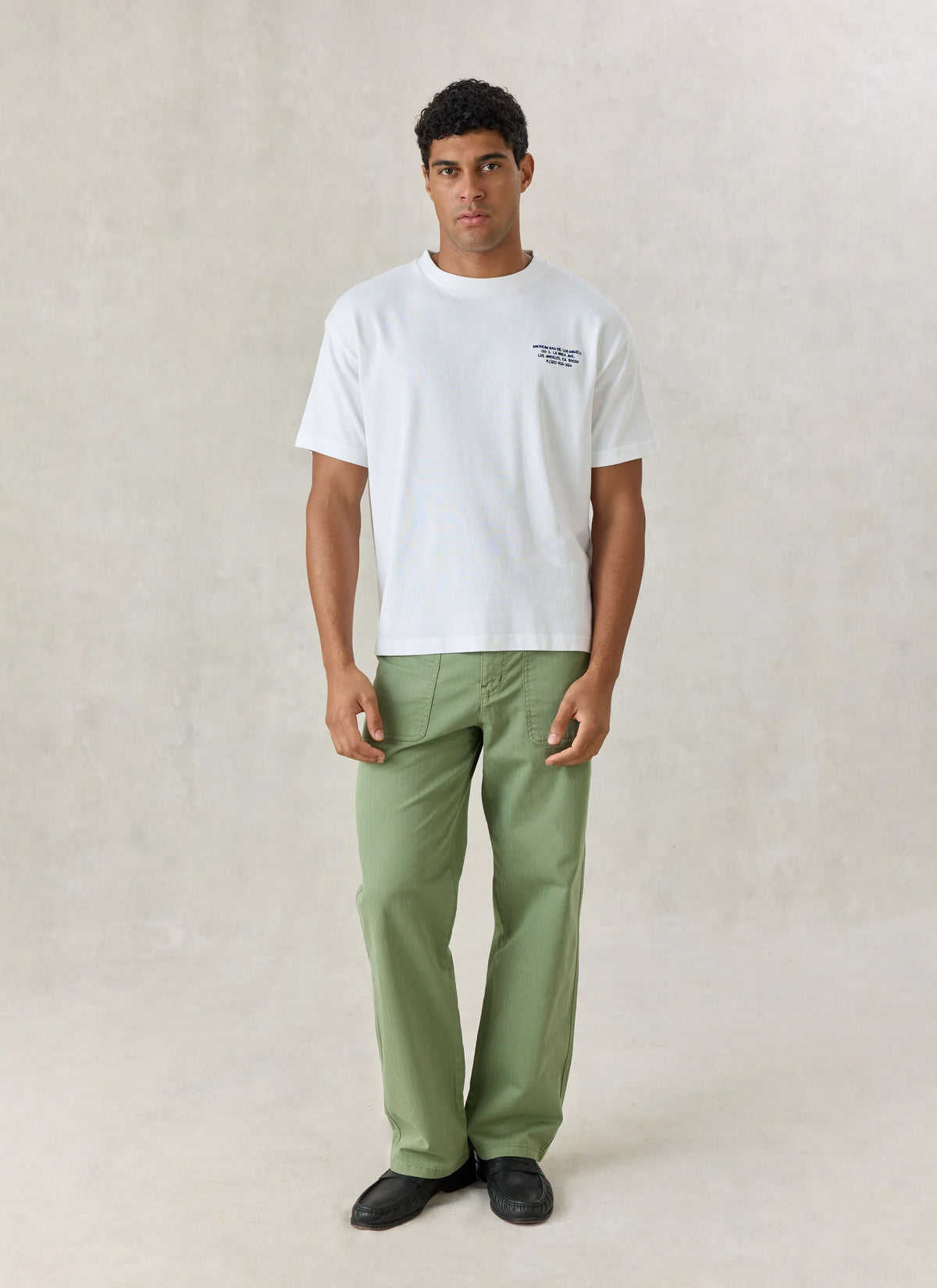 A man modeling a white crewneck t-shirt with a small dark graphic on the left chest, paired with sage green trousers and black shoes, standing against a textured beige studio backdrop.