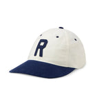 A baseball cap with an off-white crown and a navy blue brim, featuring a large embroidered navy blue letter 'R' on the front. The cap is photographed on a seamless white background.