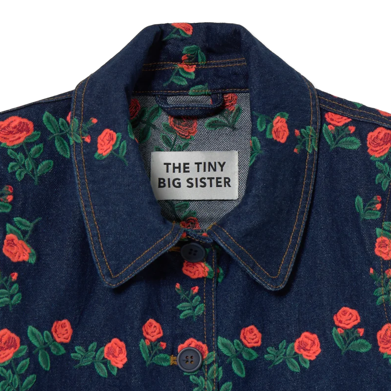 Close-up of the collar on The Tiny Big Sister Roses Grid Embroidery Denim Jacket, showing the brand label, the embroidered roses, the navy buttons and the contrast topstitching.