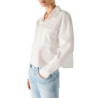 Three-quarter view of a model wearing the AMO Ruth Crop Shirt in white with light-wash jeans, one hand in her pocket, showing the chest pocket and cropped length.