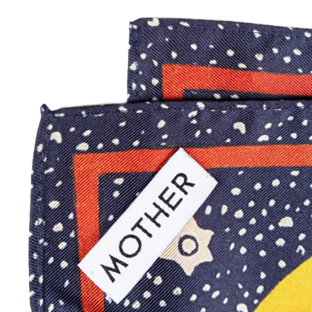 Close-up of a folded scarf with a navy blue base, white speckled pattern, orange border, and a white label reading 'MOTHER'.