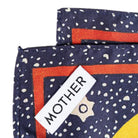 Close-up of a folded scarf with a navy blue base, white speckled pattern, orange border, and a white label reading 'MOTHER'.